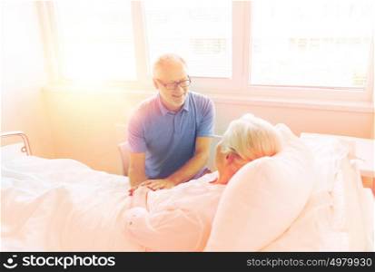 medicine, age, support, health care and people concept - happy senior man visiting and cheering his woman lying in bed at hospital ward. senior couple meeting at hospital ward