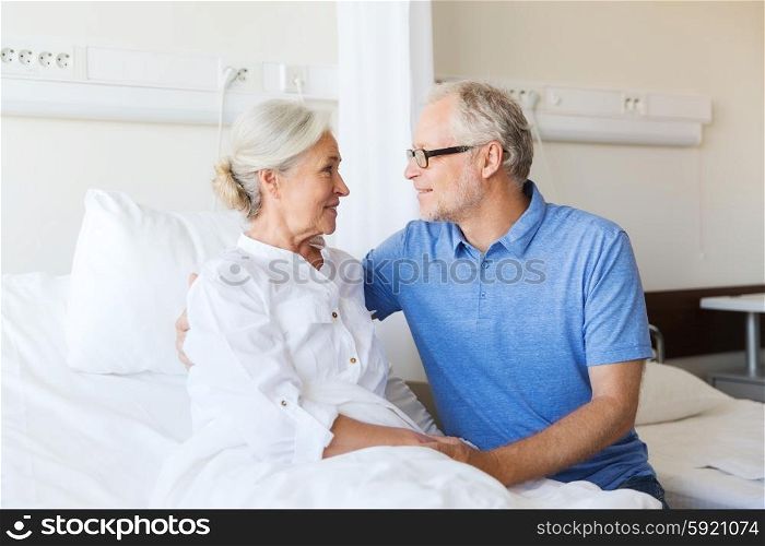 medicine, age, support, health care and people concept - happy senior man visiting and cheering his woman lying in bed at hospital ward