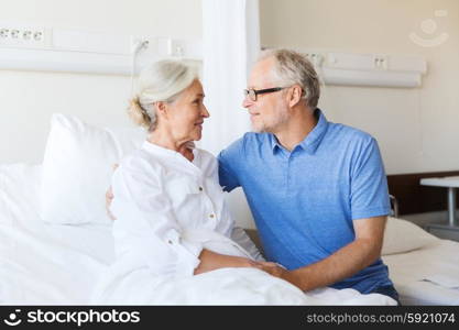 medicine, age, support, health care and people concept - happy senior man visiting and cheering his woman lying in bed at hospital ward