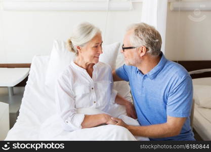 medicine, age, support, health care and people concept - happy senior man visiting and cheering his woman lying in bed at hospital ward