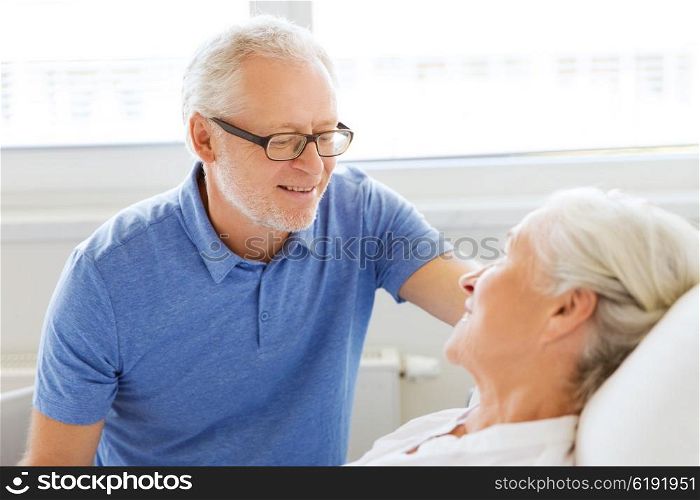 medicine, age, support, health care and people concept - happy senior man visiting and cheering his woman lying in bed at hospital ward