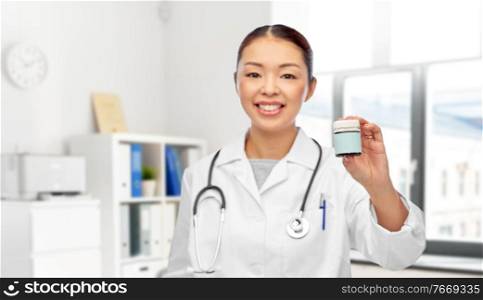medicine and healthcare concept - close up of happy smiling asian female doctor with stethoscope holding jar of pills over medical office at hospital on background. smiling female doctor holding jar of medicine