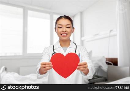 medicine, cardiology and healthcare concept - happy smiling asian female cardiologist doctor or nurse with red heart over hospital ward background. asian female doctor with red heart at hospital