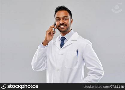 medicine, communication and healthcare concept - smiling indian male doctor or scientist in white coat with stethoscope calling on smartphone over grey background. smiling indian male doctor calling on smartphone