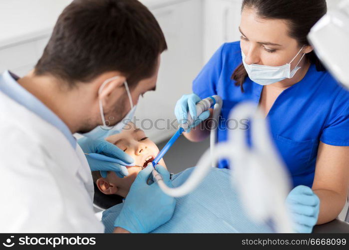 medicine, dentistry and healthcare concept - dentist and assistant with dental drill and saliva ejector treating kid patient teeth at dental clinic. dentist treating kid teeth at dental clinic. dentist treating kid teeth at dental clinic