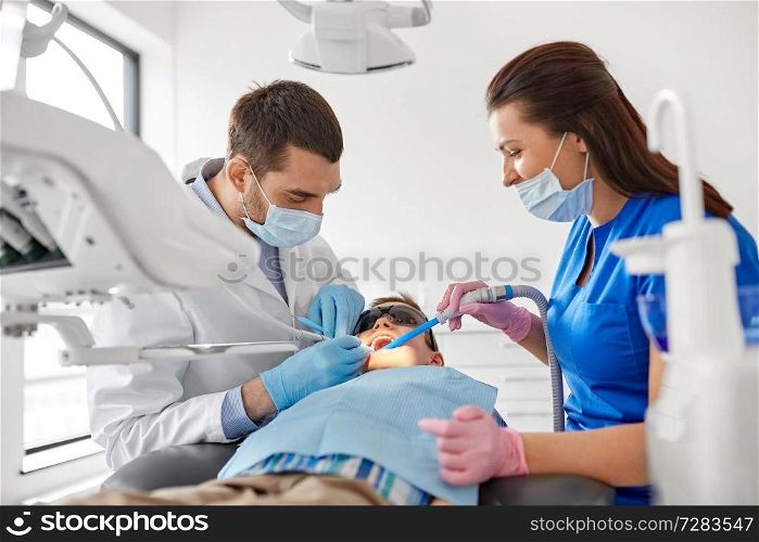 medicine, dentistry and healthcare concept - dentist and assistant with dental drill and saliva ejector treating kid patient teeth at dental clinic. dentist treating kid teeth at dental clinic