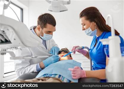 medicine, dentistry and healthcare concept - dentist and assistant with dental drill and saliva ejector treating kid patient teeth at dental clinic. dentist treating kid teeth at dental clinic