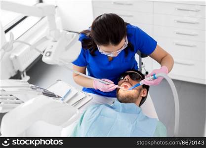 medicine, dentistry and healthcare concept - female dentist with drill and saliva ejector treating male patient teeth at dental clinic. dentist treating patient teeth at dental clinic. dentist treating patient teeth at dental clinic