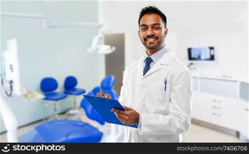 medicine, dentistry and healthcare concept - smiling indian male dentist in white coat with clipboard over dental clinic office background. indian dentist with clipboard over dental clinic
