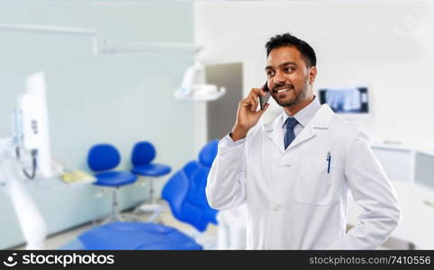 medicine, dentistry and healthcare concept - smiling indian male dentist in white coat calling on smartphone over dental clinic office background. dentist calling on smartphone at dental clinic