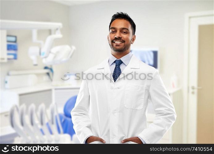 medicine, dentistry and profession concept - smiling indian male dentist in white coat over dental clinic office background. indian male dentist in white coat at dental clinic