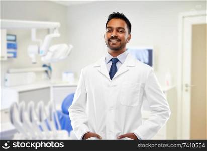medicine, dentistry and profession concept - smiling indian male dentist in white coat over dental clinic office background. indian male dentist in white coat at dental clinic