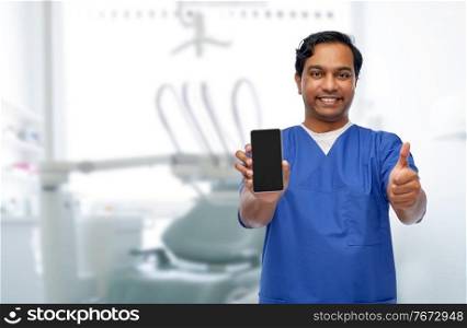 medicine, dentistry and technology concept - happy smiling indian doctor or male dentist in blue uniform with smartphone showing thumbs up over dental clinic background. doctor or dentist with phone showing thumbs up