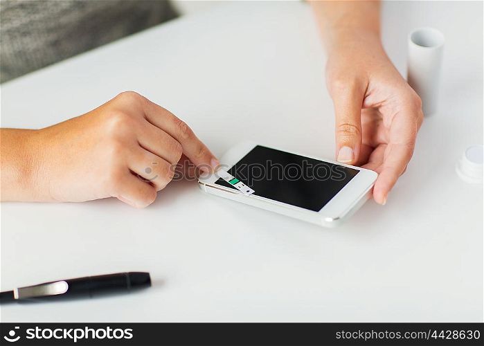 medicine, diabetes, glycemia, health care and people concept - close up of woman hands with smartphone checking blood sugar level by glucometer at home