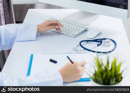 Medicine doctor&rsquo;s working on desk. Closeup of Stethoscope. Hand of Asian woman physician writing record patient information on paper note front PC computer at hospital office, Healthcare medic concept