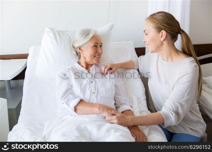 medicine, family, healthcare and people concept - daughter visiting and cheering her mother lying in bed at hospital ward. daughter visiting senior mother at hospital