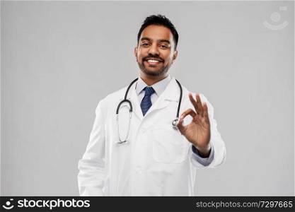 medicine, gesture and healthcare concept - smiling indian male doctor in white coat with stethoscope showing ok hand sign over grey background. smiling indian male doctor showing ok gesture