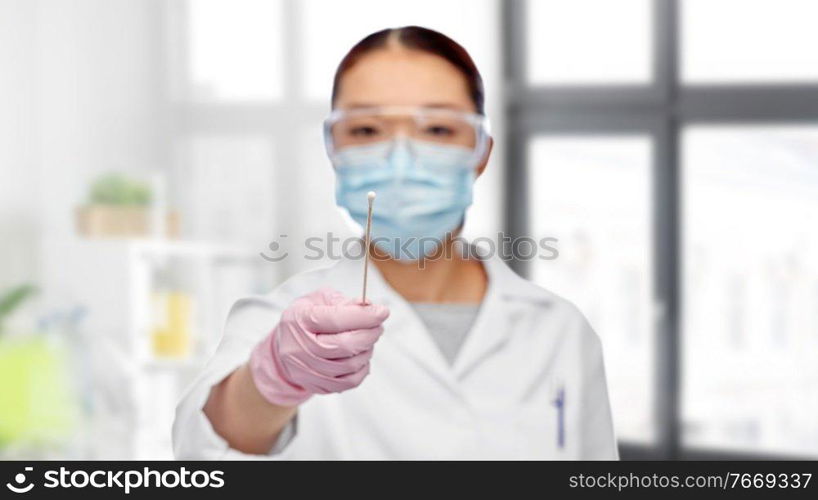 medicine, health and pandemic concept - close up of young asian female doctor or scientist in medical mask and goggles holding test cotton swab over medical office at hospital on background. asian female doctor in mask with test cotton swab