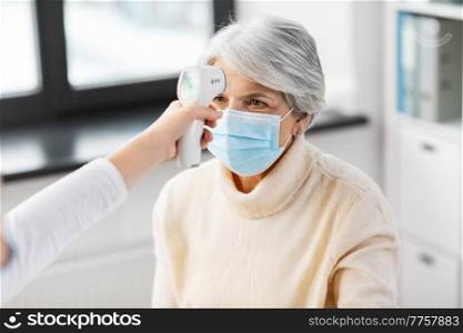 medicine, health and pandemic concept - doctor with infrared thermometer gun checking senior woman’s temperature at hospital. doctor with thermometer and woman at hospital