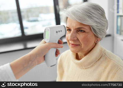 medicine, health and people concept - doctor with infrared thermometer gun checking senior woman’s temperature at hospital. doctor with thermometer and woman at hospital