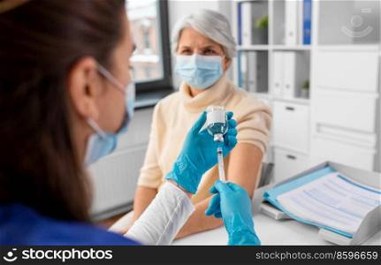 medicine, health and vaccination concept - close up of doctor or nurse filling syringe with vaccine or drug and senior woman in mask at hospital. doctor with syringe and senior woman at hospital