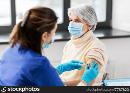 medicine, health and vaccination concept - doctor or nurse with syringe making vaccine or drug injection to senior woman in mask at hospital. nurse with syringe making injection to woman