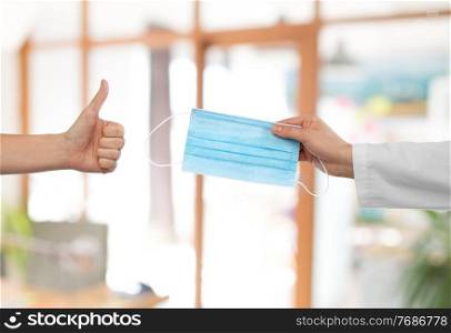 medicine, health protection and healthcare concept - hand of doctor with medical mask and patient showing thumbs up over office background. hand with mask and showing thumbs up at office