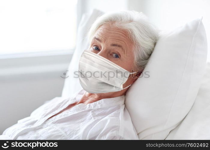 medicine, health safety and pandemic concept - senior woman patient lying in bed wearing face protective medical mask for protection from virus disease at hospital ward. old woman patient in mask lying in bed at hospital