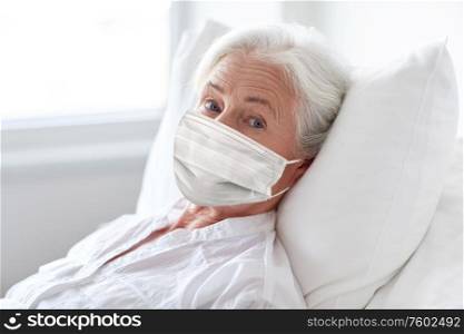 medicine, health safety and pandemic concept - senior woman patient lying in bed wearing face protective medical mask for protection from virus disease at hospital ward. old woman patient in mask lying in bed at hospital