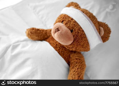 medicine, healthcare and childhood concept - ill teddy bear toy with bandaged head lying in bed. ill teddy bear toy with bandaged head lying in bed