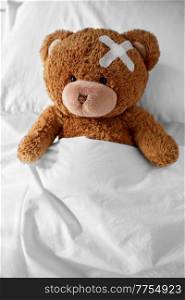 medicine, healthcare and childhood concept - ill teddy bear toy with medical patch on head lying in bed. teddy bear with medical patch on head lying in bed