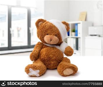 medicine, healthcare and childhood concept - teddy bear toy with bandaged head and paw over medical office at hospital background. teddy bear with bandaged head and paw at clinic