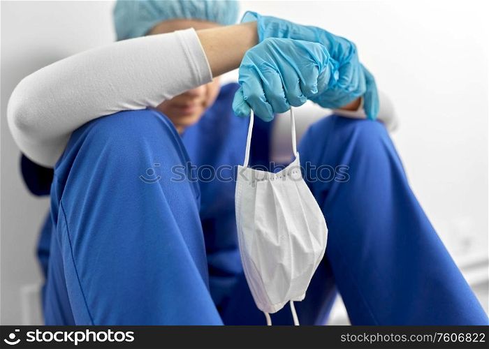 medicine, healthcare and pandemic concept - sad young female doctor or nurse holding face protective mask for protection from virus disease sitting on floor at hospital. sad doctor or nurse holding protective face mask