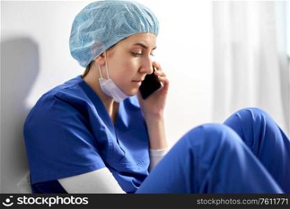medicine, healthcare and pandemic concept - sad young female doctor or nurse sitting on floor and calling on smartphone. sad doctor or nurse calling on smartphone