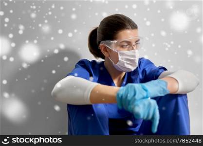 medicine, healthcare and pandemic concept - sad young female doctor or nurse wearing goggles and face protective mask for protection from virus disease sitting on floor at hospital in winter over snow. sad doctor or nurse in goggles and face mask