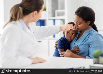 medicine, healthcare and pediatry concept - african american mother with sick son and caucasian doctor measuring baby&rsquo;s temperature by hand at clinic. mother with sick baby son and doctor at clinic