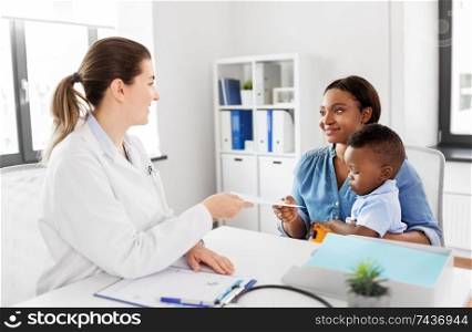 medicine, healthcare and pediatry concept - caucasian doctor giving prescription to african american mother with baby son at clinic. happy mother with baby son and doctor at clinic