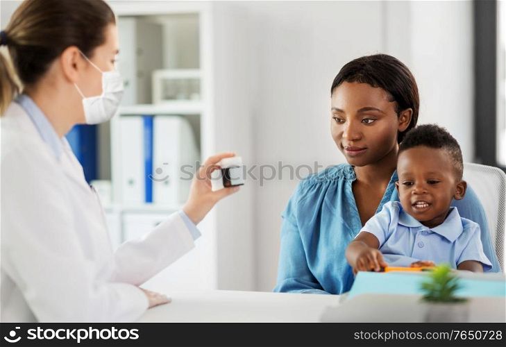 medicine, healthcare and pediatry concept - caucasian doctor wearing face protective medical mask for protection from virus disease giving medication to african american mother with baby son at clinic. doctor in mask and woman with son at clinic