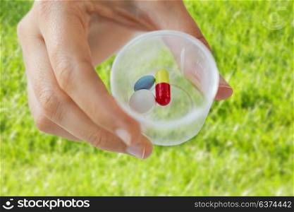 medicine, healthcare and people concept - close up of female hand holding cup with pills over grass background. close up of female hand with pills in medicine cup
