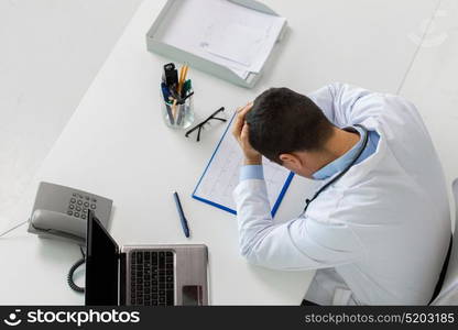 medicine, healthcare and people concept - doctor with cardiogram on clipboard and laptop computer holding to head at table in clinic. doctor with cardiogram and laptop at clinic