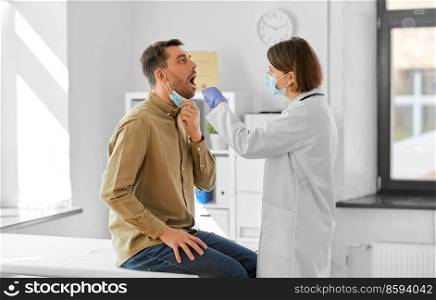 medicine, healthcare and people concept - female doctor in mask with tongue depressor checking sore throat of man patient at hospital. doctor checking male patient’s throat at hospital