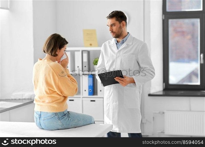 medicine, healthcare and people concept - male doctor with clipboard talking to woman patient blowing her nose with paper tissue at hospital. male doctor and woman blowing nose at hospital