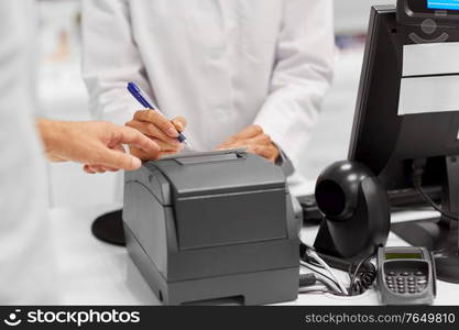 medicine, healthcare and people concept - pharmacist and customer with prescription at cash register at pharmacy. pharmacist and customer at cash register