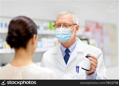 medicine, healthcare and people concept - senior apothecary wearing face protective medical mask for protection from virus disease showing to female customer at pharmacy. apothecary in mask showing drug to customer