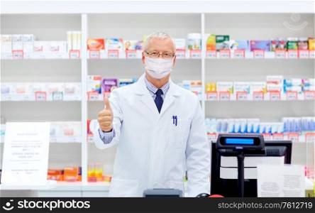 medicine, healthcare and people concept - senior apothecary wearing face protective medical mask for protection from virus disease at pharmacy cash register showing thumbs up. old apothecary in mask at pharmacy shows thumbs up