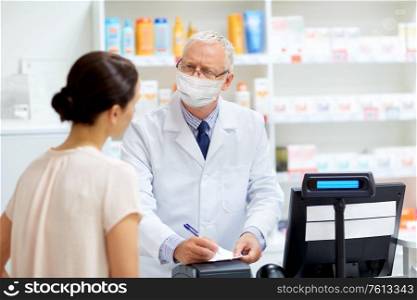 medicine, healthcare and people concept - senior apothecary wearing face protective medical mask for protection from virus disease writing prescription for female customer at pharmacy. senior apothecary in mask and customer at pharmacy