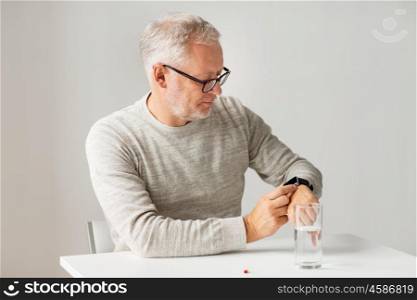 medicine, healthcare and people concept - senior man with glass of water water and pill looking at wristwatch or smartwatch with health application at home