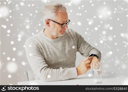 medicine, healthcare and people concept - senior man with glass of water water and pill looking at wristwatch or smartwatch with health application at home over snow. senior man with water and pill looking at watch