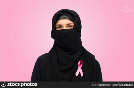 medicine, healthcare and people concept - smiling muslim female doctor wearing hijab and white coat with pink breast cancer awareness ribbon. muslim woman with breast cancer awareness ribbon