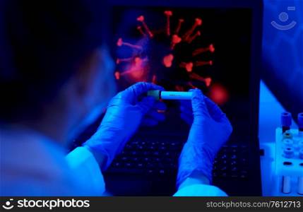 medicine, healthcare and science concept - close up of hand in protective medical glove holding beaker with virus positive blood test over coronavirus on laptop computer screen at laboratory. hand holding beaker with coronavirus blood test